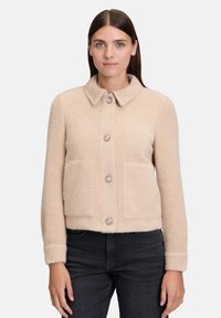 Kobieta nosząca beżową, strukturalną wełnianą kurtk ę z kołnierzem, czterema guzikami i dwiema bocznymi kieszeniami, w połączeniu z czarnymi dżinsami, stojąca na tle białym.