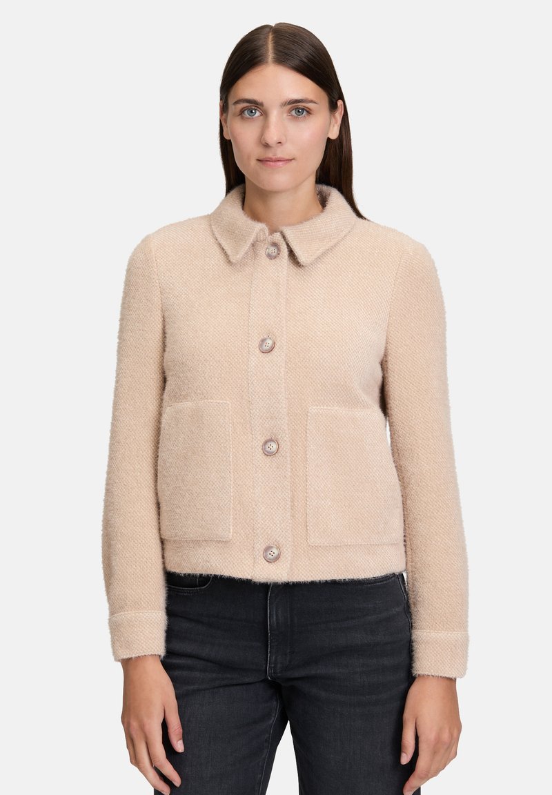 Kobieta nosząca beżową, strukturalną wełnianą kurtkę z kołnierzem, czterema guzikami i dwiema bocznymi kieszeniami, w połączeniu z czarnymi dżinsami, stojąca na tle białym.