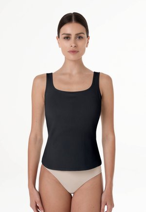 Donna con capelli scuri che indossa un top nero a coste senza maniche e slip beige a vita alta, in piedi davanti a uno sfondo bianco semplice.