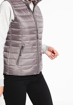 Persona con un chaleco sin mangas acolchado de color gris claro sobre una camisa gris de manga larga y pantalones negros, con cremallera y bolsillo lateral visible.