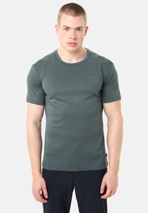 Junger Mann mit kurzen Haaren, der ein eng anliegendes dunkelgrünes kurzärmeliges T-Shirt und schwarze Hosen trägt, steht vor einem schlichten hellen Hintergrund.