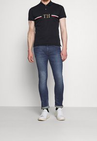 Černé polo tričko se zlatým logem "TH", horizontální barevné pruhy. Nosené s tmavě modrými skinny džínami a bílými teniskami, s ohrnutými rukávy.