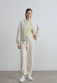 Ελαφρύ bomber τζάκετ με καπιτονέ υφή σε ανοιχτό γκρι χρώμα, πάνω από παστέλ κίτρινο πουκάμισο και εκρού φόρμες. Λευκά αθλητικά παπούτσια ολοκληρώνουν την εμφάνιση. Λείες υφές.