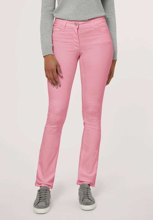 Partie inférieure du corps d'une personne portant un jean slim rose clair, un pull gris et des baskets grises, debout sur un fond neutre.