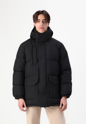 Black puffer jacket with a high collar, visible stitching, and two large front pockets. Elastic cuffs and a zip closure are featured.