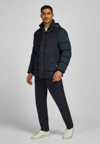 Marineblauwe pufferjack met een matte afwerking, ritszakken en een afneembare capuchon; gedragen met bijpassende marineblauwe broeken en witte sneakers.