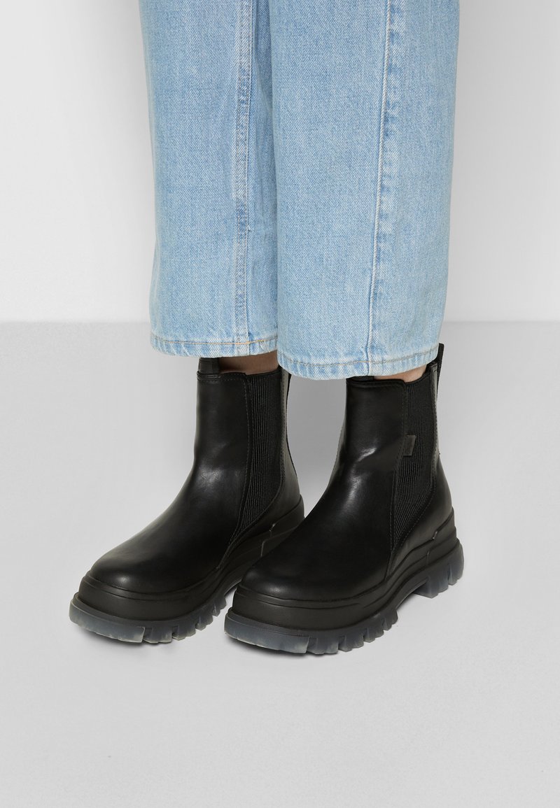 Schwarze Leder-Stiefeletten mit elastischen Seiteneinsätzen, einer strukturierten Oberfläche und robusten grauen Gummisohlen. Kombiniert mit hellblauen, cropped Jeans.