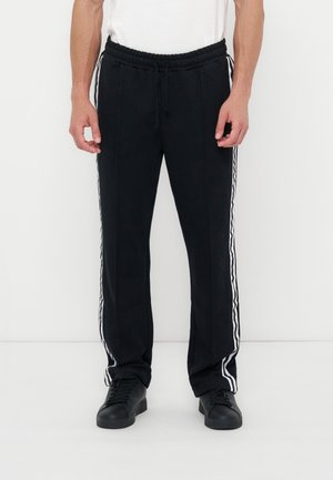 Personne portant un pantalon de sport noir avec des bandes blanches sur les côtés et des baskets noires, debout devant un fond blanc.