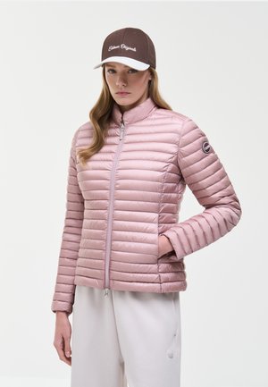 Ung kvinde iført en lyserød quiltet jakke med lynlås og en brun og hvid baseballkasket, stående med den ene hånd i lommen.