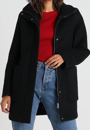 Cappotto in misto lana nero con zip frontale, grandi tasche a toppa e cappuccio. Indossato sopra un top rosso a coste e jeans in denim azzurro chiaro.