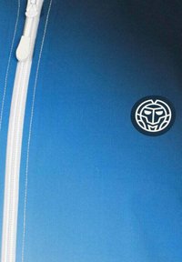 Chaqueta azul con cremallera blanca, costuras blancas y un logo circular con un diseño de cara en la parte inferior derecha. Textura suave.