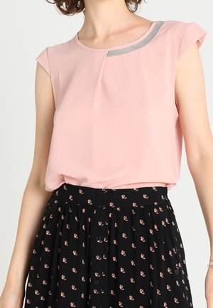 Femme portant un chemisier sans manches rose clair avec un accent gris près de l'encolure et une jupe noire avec de petits motifs floraux roses.