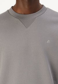 Grauer Sweatshirt mit geripptem Rundhalsausschnitt und V-Einsatzdetail. Mit kleinem, hellgrauen Logo auf der Brust. Hergestellt aus weichem Textil.