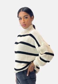 Stickad tröja i krämfärg med svarta horisontella ränder, med hög krage och lösa ärmar. Kombinerad med mörka jeans.