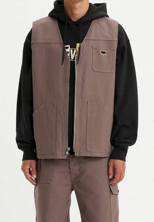 Mężczyzna ubrany w fioletowawy kamizelkę roboczą z przednimi kieszeniami, nałożoną na czarną bluzę z kapturem oraz dopasowane fioletowawa spodnie cargo, stojący na jednolitym tle.