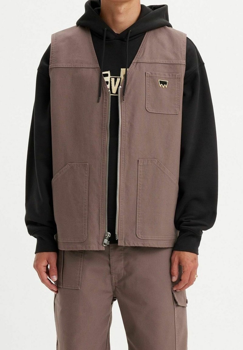 Homme portant un gilet utilitaire mauve avec poches avant sur un sweat à capuche noir et un pantalon cargo mauve assorti, debout devant un fond uni.
