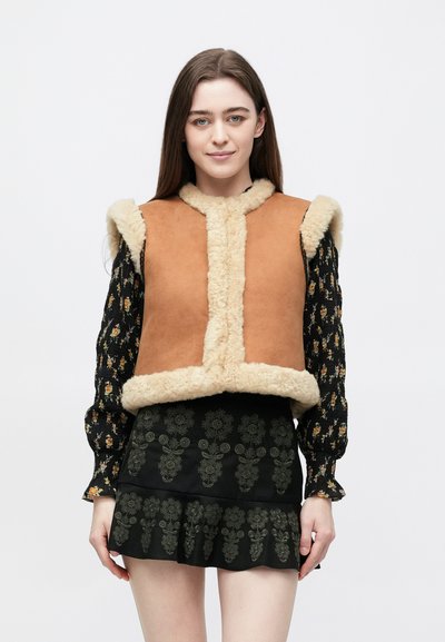 Brun ruskindsvest med beige shearling-krave og kant, med en tætsiddende pasform. Båret over en sort bluse og nederdel med blomstermønster.