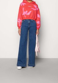 Sudadera corta roja con patrón abstracto en rosa, combinada con jeans de denim azul de pierna ancha. La modelo sostiene un bolso rosa claro y usa zapatillas blancas.