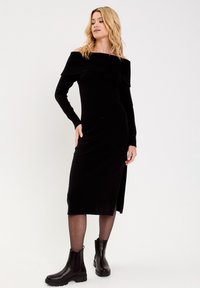 Robe en tricot noire à épaules dénudées avec encolure côtelée, manches longues et fente sur le côté. Portée avec des bottines noires. Texture lisse, design ajusté.