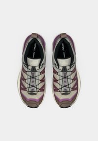 Παπούτσια για ορεινό τρέξιμο σε γκρι με μωβ λεπτομέρειες. Διαθέτουν υφαντό πλέγμα στην επάνω πλευρά, μαύρα κορδόνια, λαστιχένια σόλα και λογότυπο στη γλώσσα.