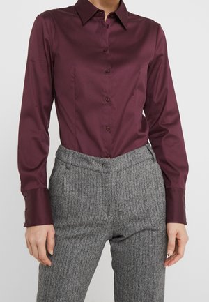 Nainen pukeutuneena istuvaan pitkähihaiseen burgundinpunaisen sävyiseen napitettuun paitaan, joka on puettu korkeavyötäröisten harmaiden kalanruotohousujen sisään.