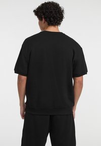 Sort bomulds oversized t-shirt med korte ærmer, rund hals og ribbet kant, set fra ryggen, glat tekstur uden synlige mønstre.