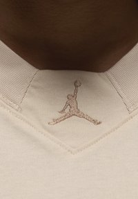 Tessuto color crema con colletto testurizzato. Presenta un logo Jumpman ricamato marrone chiaro all'altezza del collo, che simboleggia l'abbigliamento sportivo.