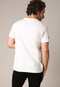 T-shirt en coton blanc avec un col rond simple et des manches courtes, arborant un petit logo noir sur la manche supérieure gauche.