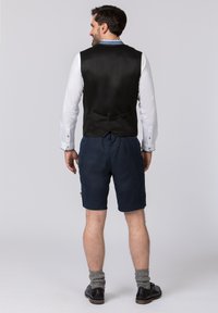 Schwarze Satin-Weste mit einem Gurt hinten, getragen über einem weißen Hemd. Kombiniert mit dunkelblauen Shorts und grauen Kniestrümpfen. Schwarze Schuhe runden den Look ab.