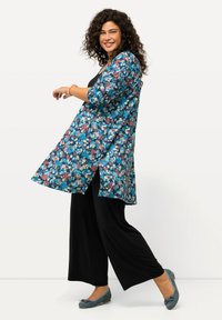 Cárdigan largo floral en azul, con flores rosas y blancas, y aberturas laterales. Combinado con pantalones anchos negros y zapatos planos en color verde azulado.