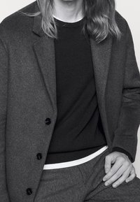 Grå ullkavaj med svarta knappar över en svart tröja, med en vit fåll synlig vid midjan. Grå byxor fulländar outfiten.