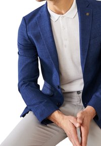 Niebieska teksturowana marynarka na białej koszulce polo, zestawiona z jasnoszarymi spodniami. Charakterystycznym elementem jest przypinka z logo na klapie marynarki.