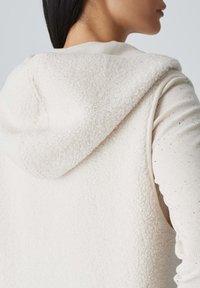 Fleece-Weste in Off-White mit einer weichen, strukturierten Kapuze, darüber eine leichte Crewneck-Oberbekleidung mit dezentem, gesprenkeltem Muster.