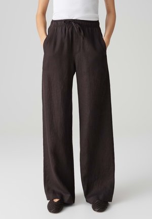 Person wearing black wide-leg pants with drawstring waist, white sleeveless top, and black woven slip-on shoes, standing hands in pockets.