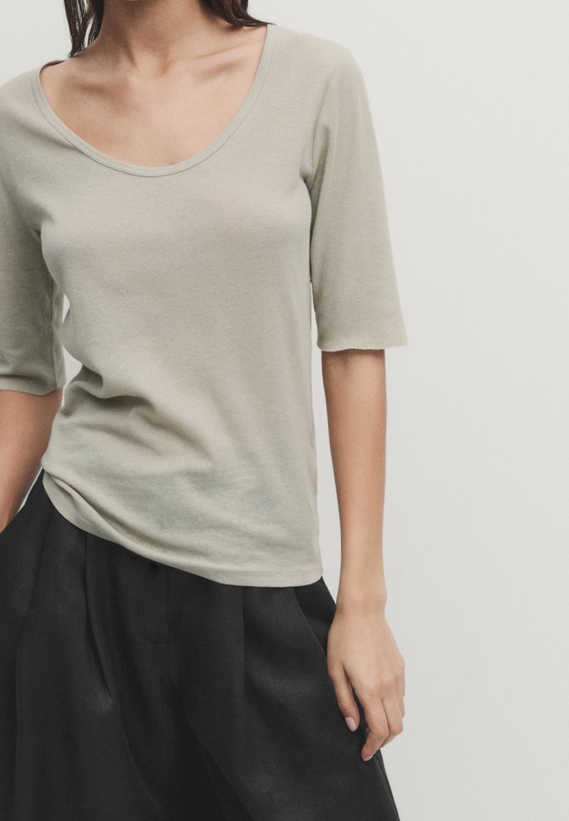 Femme portant un haut gris clair à encolure dégagée avec manches aux coudes et un pantalon noir ample sur un fond uni.