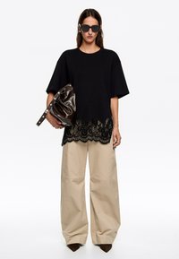 Must mustriga must-särgid, millel on pitsäär, koos khaki laiade pükstega. Modell hoiab käes läikivat pruuni kotti ja kannab tumedaid päikeseprille.