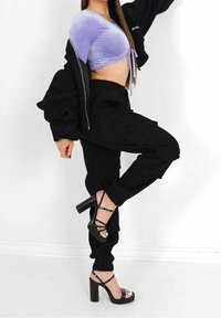 Vrouw tilt één been op, gekleed in een paars cropped top, zwarte cargobroek, zwarte sandalen met hoge hakken en een gedeeltelijk gedragen zwarte jas.