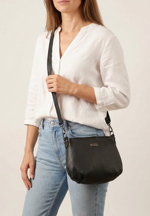 Kvinde iført hvid langærmet skjorte og lyseblå jeans, der holder en sort læder crossbody-taske med justerbar rem på skulderen.