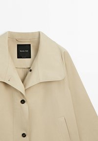 Beżowy lekki płaszcz z szerokim kołnierzem, czarnymi guzikami, boczną kieszenią i metką Massimo Dutti wewnątrz kołnierza.