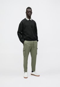 Άνδρας που στέκεται με τα χέρια στις τσέπες, φοράει μαύρο φούτερ, γκρι πουκάμισο με γιακά, χακί παντελόνι cargo και λευκά αθλητικά παπούτσια σε απλό φόντο.