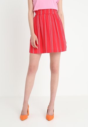Jupe rouge avec des rayures verticales blanches, taille élastique, longueur au-dessus du genou, associée à des chaussures à talons hauts orange.