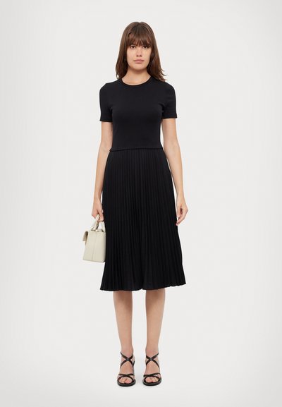 Schwarzes Kleid mit enger Oberseite und plissiertem Rock. Kombiniert mit schwarzen Riemchensandalen und einer kleinen Handtasche in Creme. Glatte Stoffoberfläche.