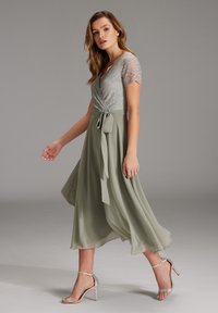 Abito avvolgente con maniche corte in pizzo, gonna in chiffon verde chiaro, vita aderente con dettaglio fiocco annodato, abbinato a sandali argentati con cinturini.