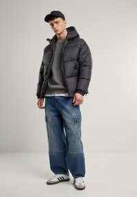 Black puffer jacket with a hood, gray knit sweater, loose blue cargo jeans, and white athletic shoes with black stripes. Plain background.