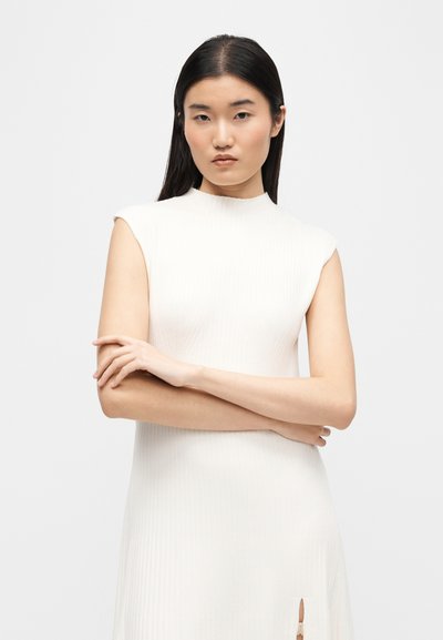 Femme aux longs cheveux noirs portant une robe blanche côtelée sans manches, bras croisés, sur un fond gris clair uni.