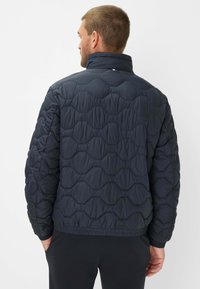 Giacca imbottita trapuntata navy con colletto alto, polsini ed orlo elastici. Il motivo a onde prominenti sul tessuto aggiunge trama e interesse visivo.