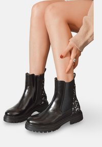 Bottines en cuir noir avec une finition brillante, panneaux latéraux élastiques et un accent en tissu à motif à l'arrière. Semelle en caoutchouc texturée.
