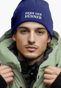 Blue knitted beanie with a rectangular patch reading "HERR DER HÜHNER." The model wears a green jacket with a black interior, showcasing a layered look.