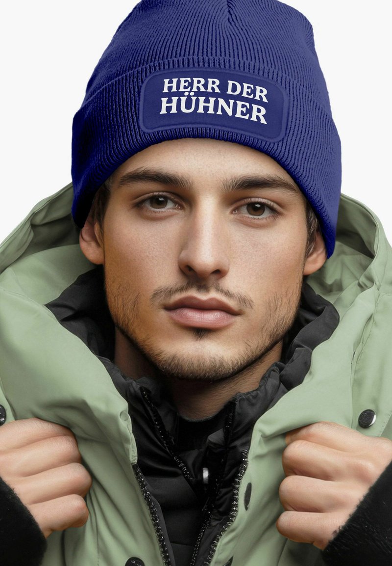 Blue knitted beanie with a rectangular patch reading "HERR DER HÜHNER." The model wears a green jacket with a black interior, showcasing a layered look.