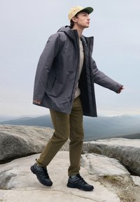 Veste imperméable grise avec capuche, pull beige, pantalon vert olive et chaussures noires. Debout sur des rochers de granit avec vue sur la montagne.